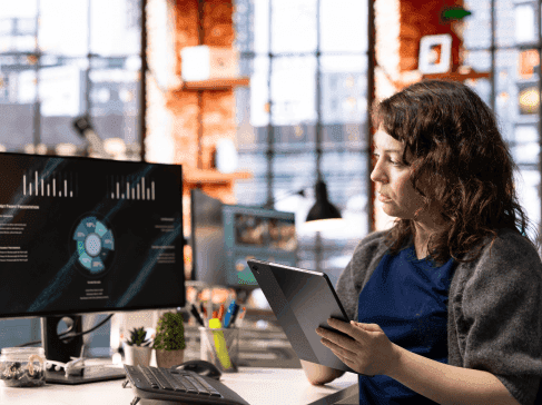 Žena sedí před počítačem s tabletem v ruce