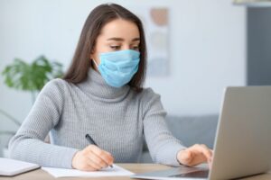 Mladá žena sedící u notebooku s rouškou během koronaviru